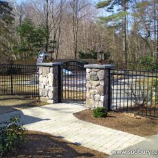 Black aluminum fence with a matching gate and stone pillars along a paved walkway, surrounded by trees and landscaped grounds. Sudbury Cedar Fence Co