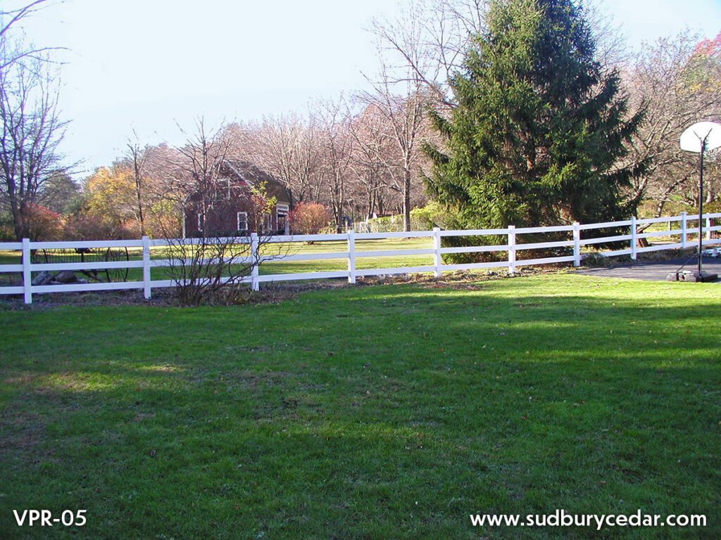 White ranch rail fence running along a green lawn with trees and a rural property in the background. Sudbury Cedar Fence Co, Bolton MA