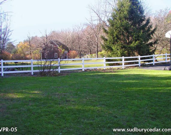 White ranch rail fence running along a green lawn with trees and a rural property in the background. Sudbury Cedar Fence Co, Bolton MA