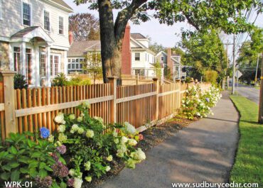 Wood picket fence along a sidewalk with flowering shrubs and traditional homes, highlighting a classic fence design that enhances curb appeal. Sudbury Cedar Fence Co. Concord, MA.