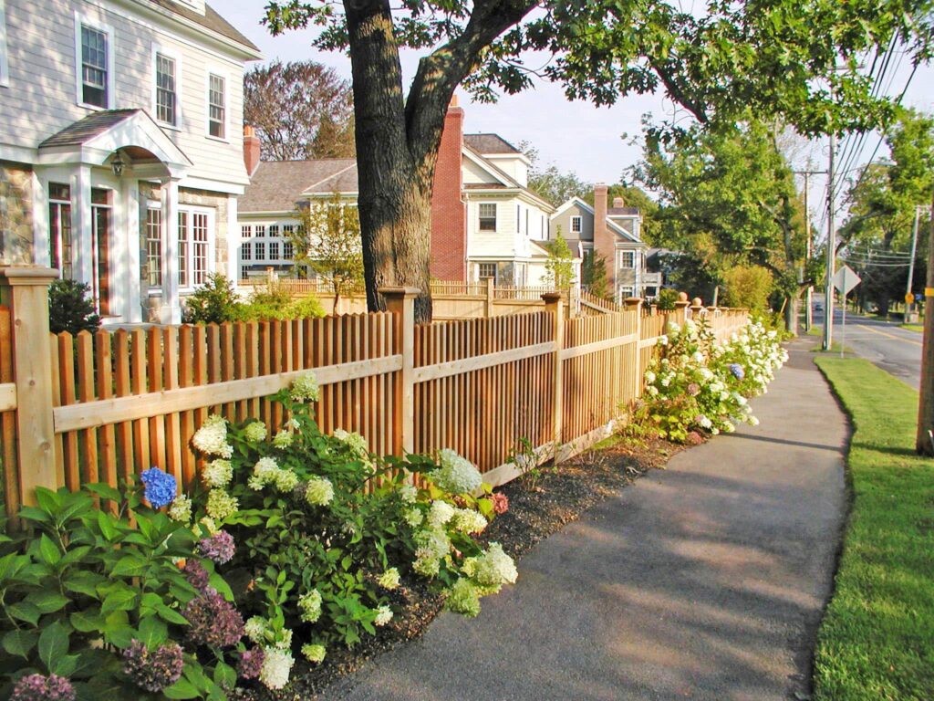 Wood picket fence along a sidewalk with flowering shrubs and traditional homes, highlighting a classic fence design that enhances curb appeal. Sudbury Cedar Fence Co, in Concord MA
