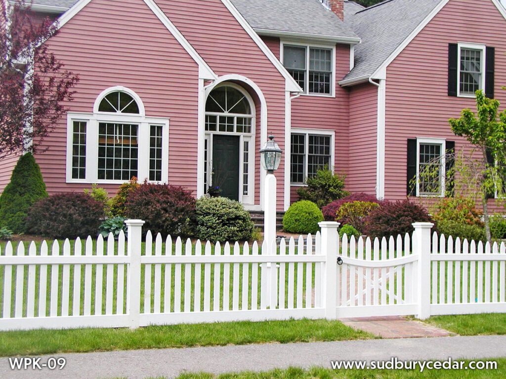 White picket fence with a gate in front of a pink colonial house with black shutters, landscaped shrubs, and a green lawn. Sudbury, MA