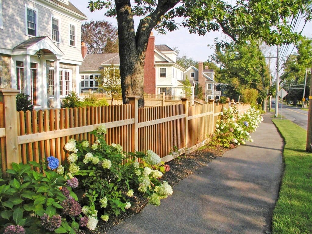 Cedar picket fence along a sidewalk with flowering shrubs and traditional homes, highlighting a classic fence design that enhances curb appeal. Sudbury Cedar Fence Co, in Concord MA