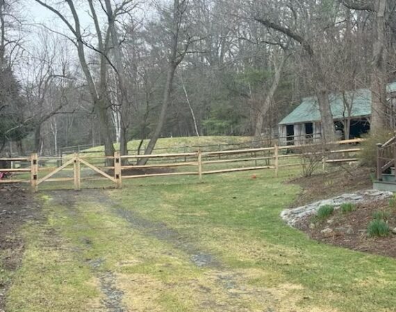 Rustic cedar post and rail fence with protective wire mesh installed on a residential property in Stow, Massachusetts.