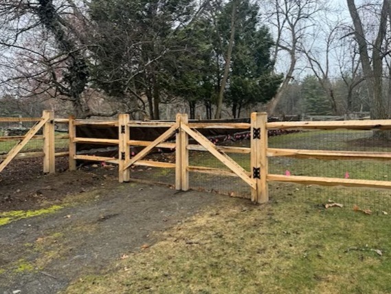 rustic cedar split-rail gate with black wire mesh backing and heavy-duty hinges, installed on a residential property in Stow, MA.