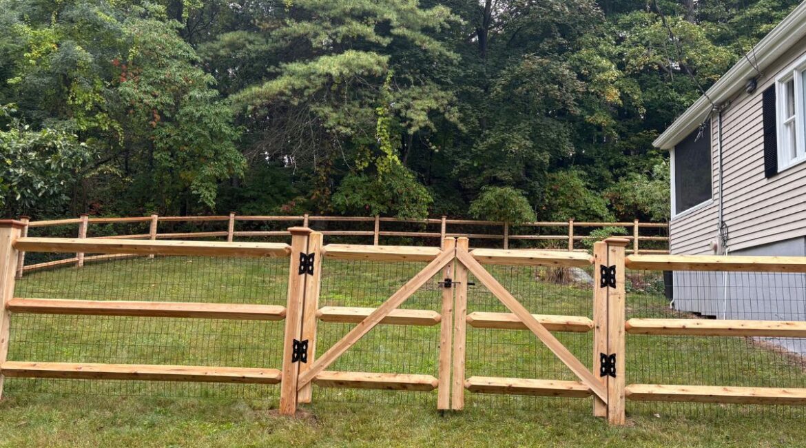 Cedar post and rail dog fence with black wire mesh and matching walk gate installed in a backyard in Sudbury, Massachusetts, providing a secure enclosure while preserving open views.
