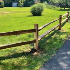 Split rail post and rail fence installed along a paved driveway in Boxborough, Massachusetts, bordering a large open lawn with ornamental grasses and mature landscaping.