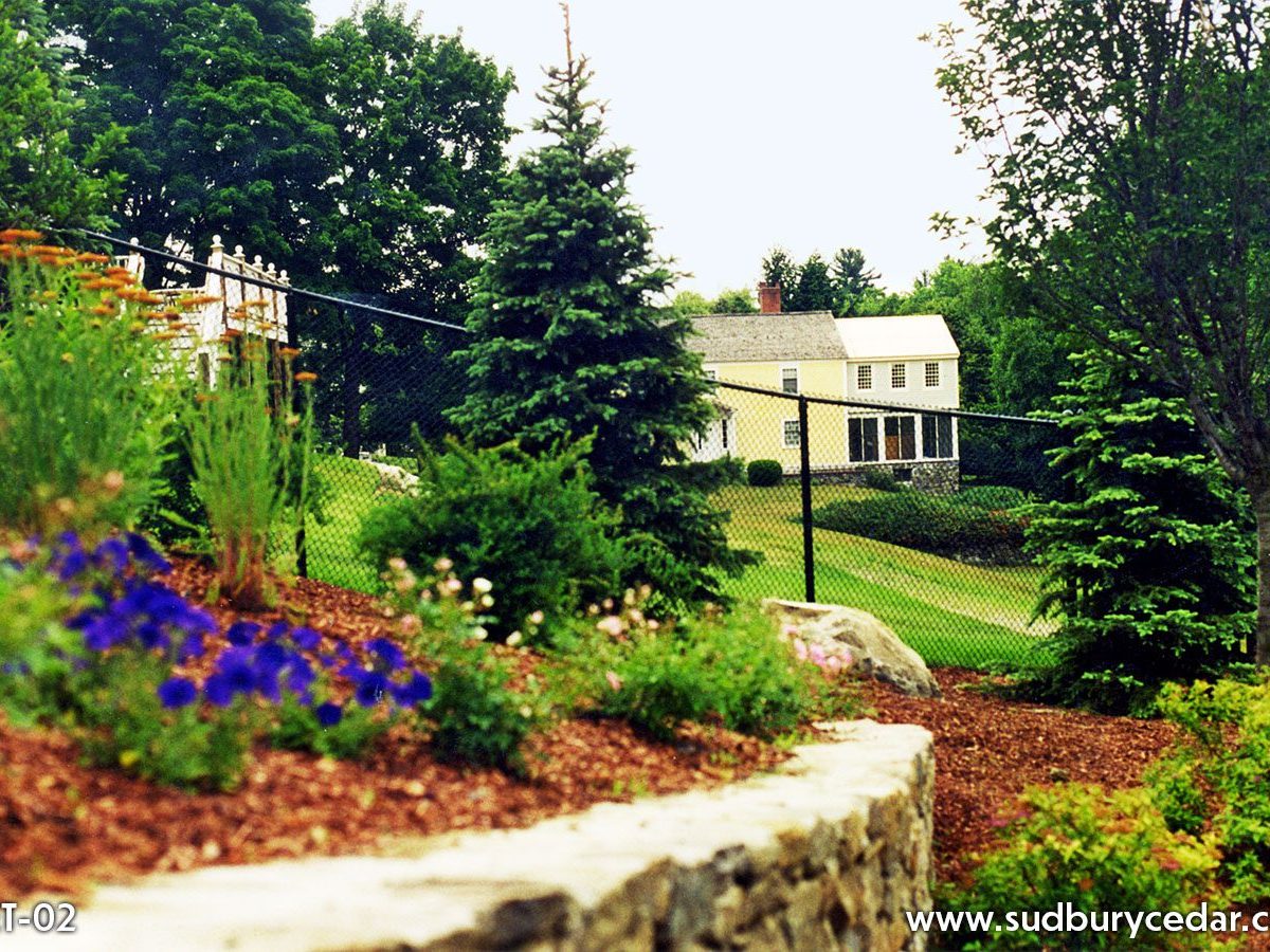 Black chain link fence installed along landscaped retaining wall in a wooded Massachusetts backyard with gardens, trees, and open lawn views.
