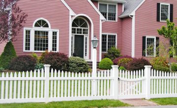 White picket fence with a gate in front of a pink colonial house with black shutters, landscaped shrubs, and a green lawn. Sudbury, MA